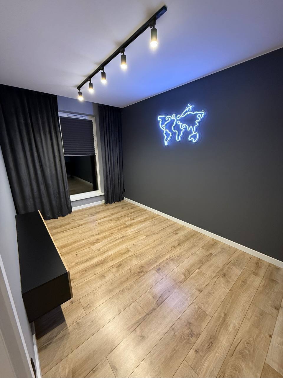Pokój po remoncie z drewnianą podłogą, szarą ścianą z neonową mapą świata i czarnymi zasłonami. Oświetlenie sufitowe punktowe. Minimalistyczny styl.
