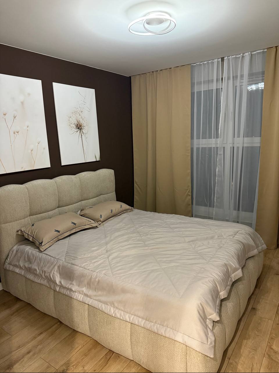 Beżowe łóżko z zagłówkiem i poduszkami ze wzorem ważek w sypialni z brązową ścianą, obrazami i zasłonami w odcieniach beżu. Parkiet na podłodze.