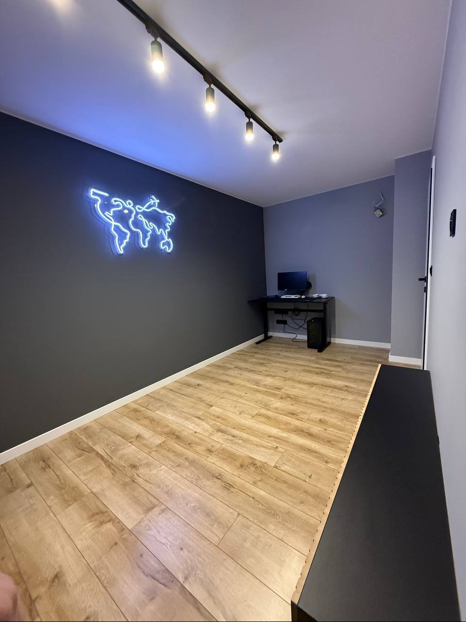 Pokój z drewnianą podłogą, szarą ścianą z neonową mapą świata i czarnym biurkiem. Oświetlenie sufitowe punktowe. Minimalistyczny design wnętrza.