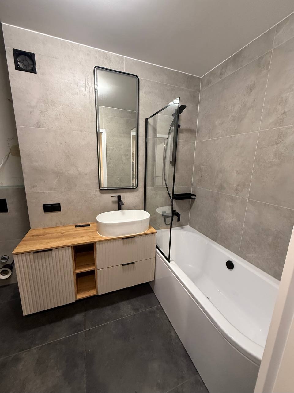 Nowoczesna łazienka z szarymi płytkami, drewnianą szafką pod umywalką i czarnymi akcentami: rama lustra, bateria, kabina prysznicowa. Minimalistyczny design.