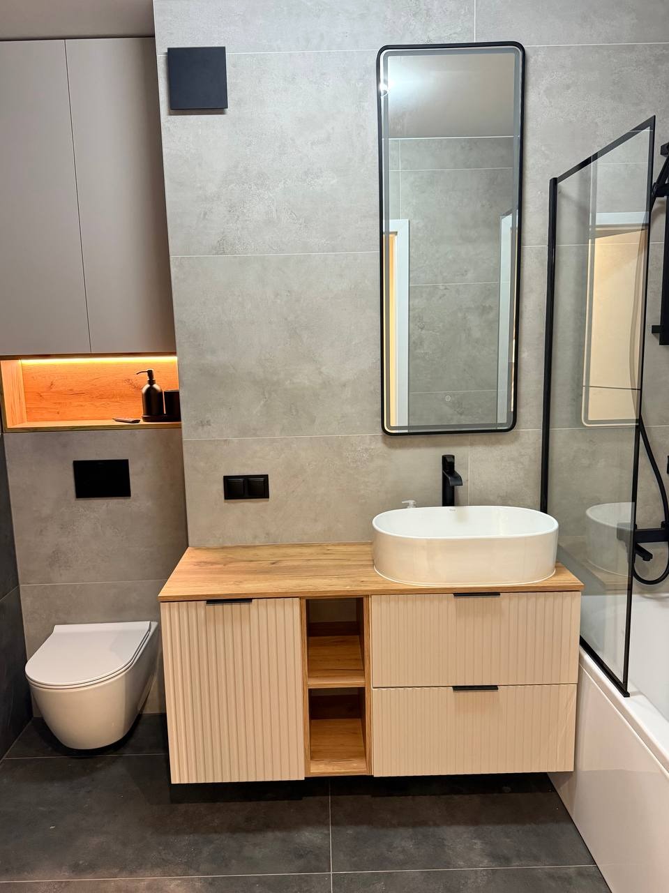 Nowoczesna łazienka z owalną umywalką na szafce z ryflowanymi frontami, podświetlana wnęka i szklana kabina prysznicowa. Szare płytki i czarne akcenty.