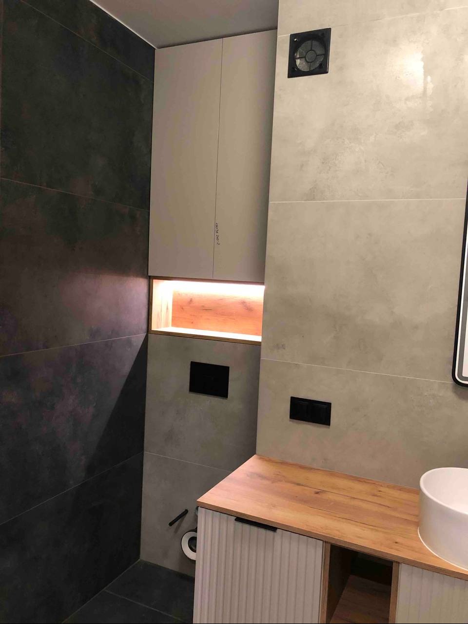 Nowoczesna łazienka z szarą glazurą, drewnianym blatem i podświetlaną wnęką. Minimalistyczny design z ukrytą spłuczką i wentylatorem.