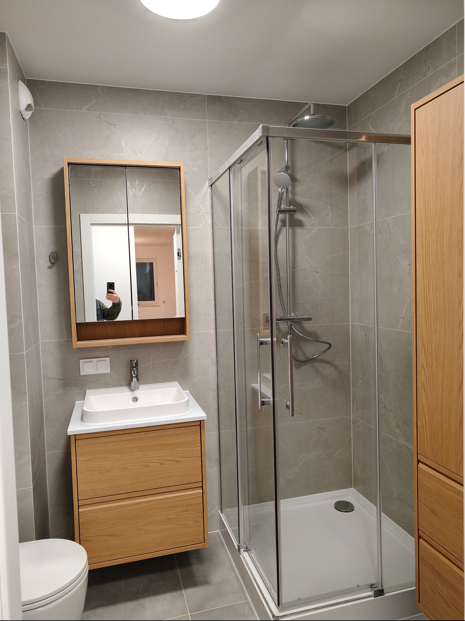 Nowoczesna łazienka z kabiną prysznicową, umywalką z szafką z jasnego drewna i lustrem. Szare płytki imitujące marmur na ścianach i podłodze. Widoczne odbicie osoby w lustrze.