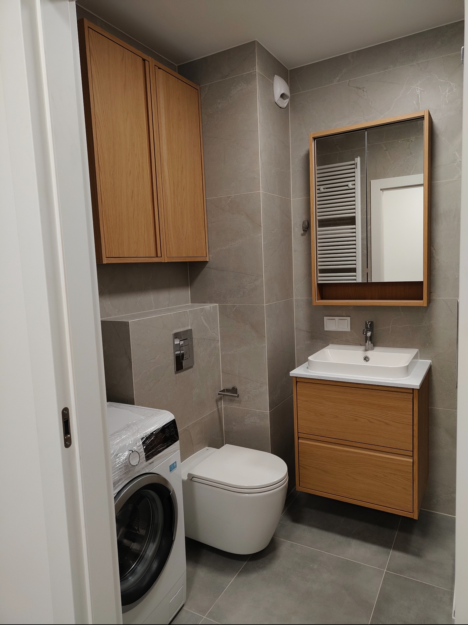Nowoczesna łazienka w szarościach z drewnianymi akcentami: szafka nad toaletą, umywalka z szafką i lustro. Pralka zabezpieczona folią. Minimalistyczny design.