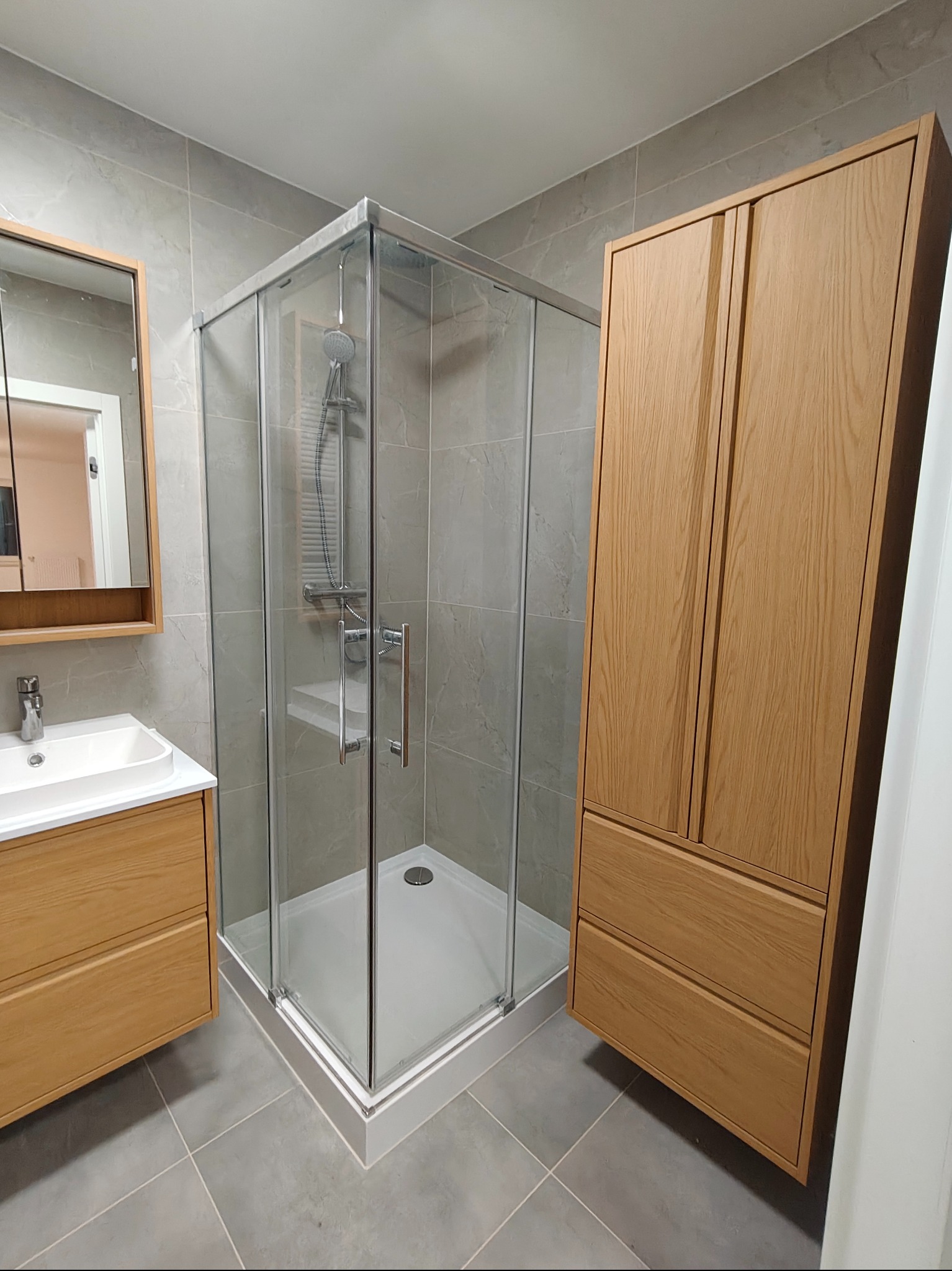 Jasna łazienka po remoncie: kabina prysznicowa z brodzikiem, umywalka z szafką i wysoka szafa z drewna. Szare płytki na ścianach i podłodze.