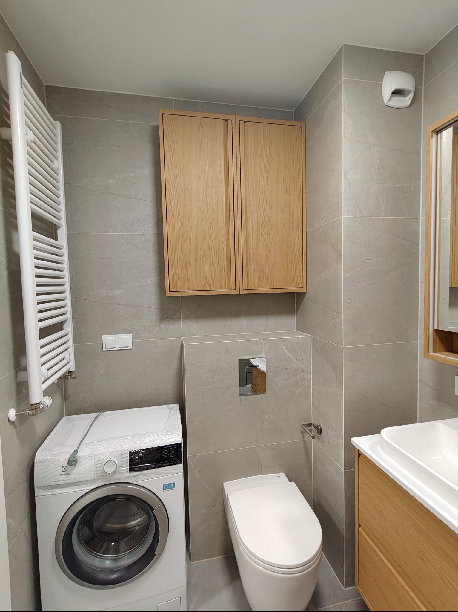 Nowoczesna łazienka w szarościach z drewnianymi akcentami: szafka nad toaletą, umywalka z szafką, grzejnik drabinkowy i pralka w folii ochronnej. Minimalistyczny design.
