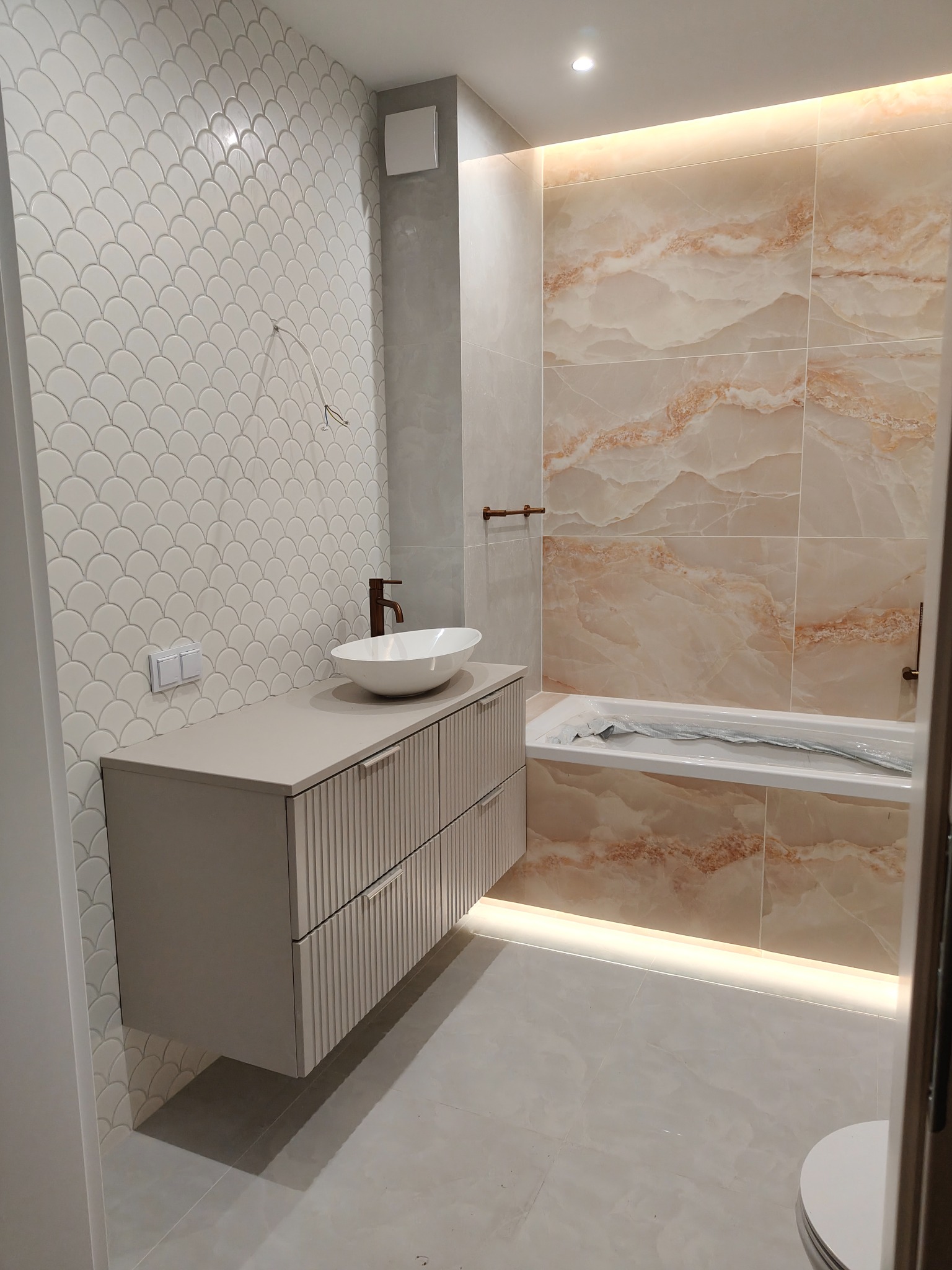 Wykończona łazienka z umywalką nablatową na szafce z frezowanymi frontami, mozaiką na ścianie i płytkami imitującymi marmur przy wannie. Oświetlenie LED.