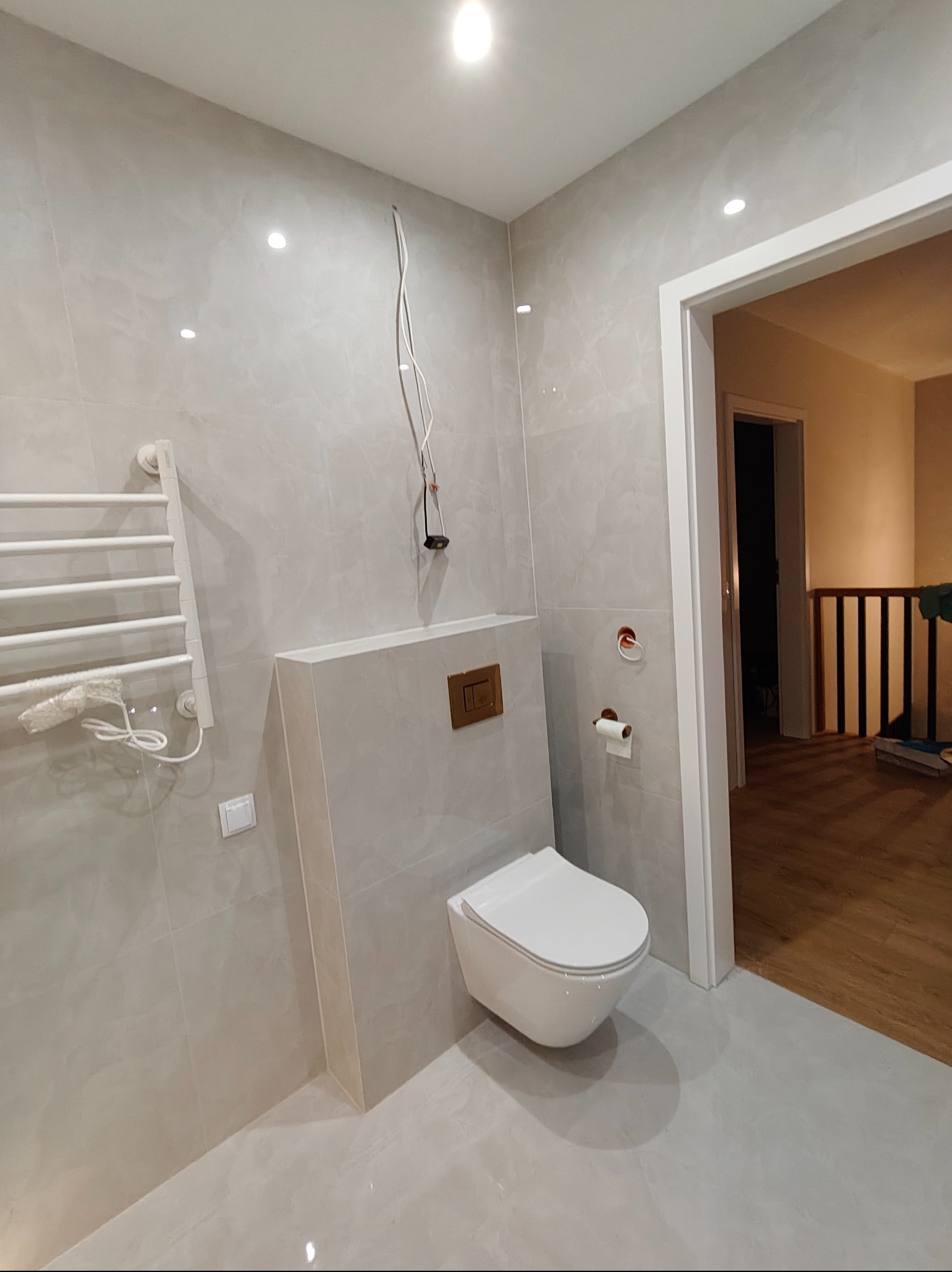 Jasna łazienka po remoncie: wisząca toaleta, grzejnik drabinkowy z ręcznikiem, marmurowe płytki i widok na drewniane schody przez otwarte drzwi.