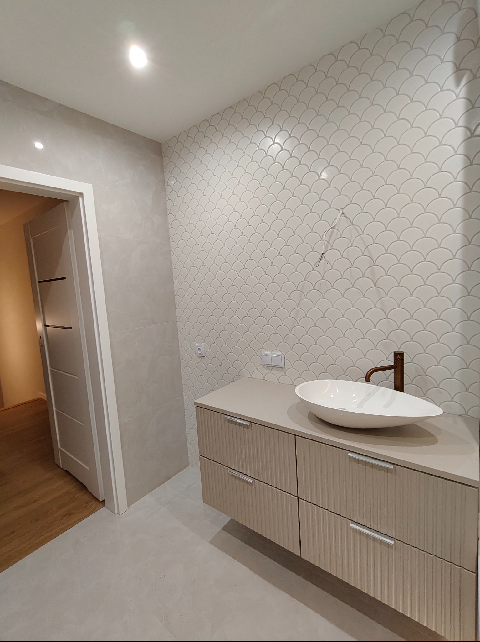 Wykończona łazienka z umywalką nablatową, baterią w kolorze miedzi i ścianą wyłożoną płytkami w kształcie rybiej łuski. Widoczna podłoga i drzwi do innego pomieszczenia.