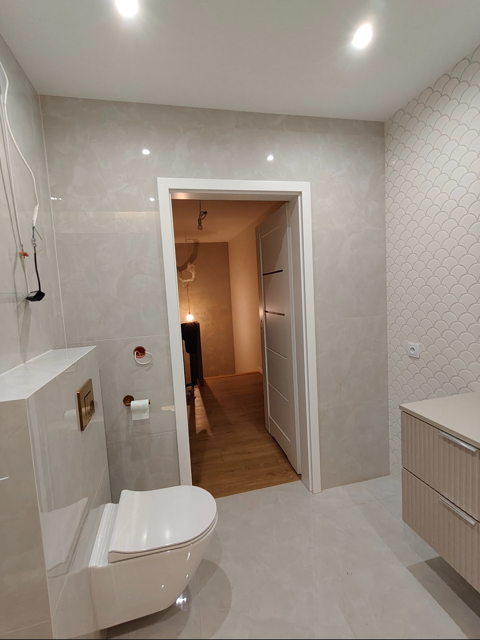 Wykończona łazienka w jasnych odcieniach szarości z wiszącą toaletą, umywalką z szafką i dekoracyjną mozaiką na ścianie. Widoczne elementy instalacji elektrycznej.