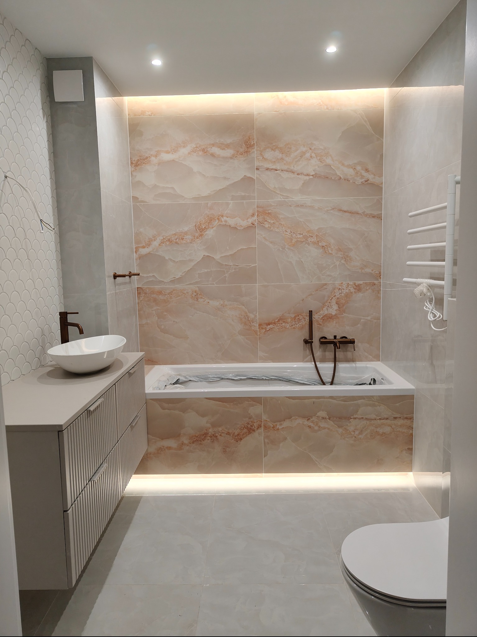 Nowoczesna łazienka z marmurowymi płytkami, podświetlaną wanną i umywalką na szafce z ryflowanymi frontami. Białe płytki w kształcie rybiej łuski na ścianie.