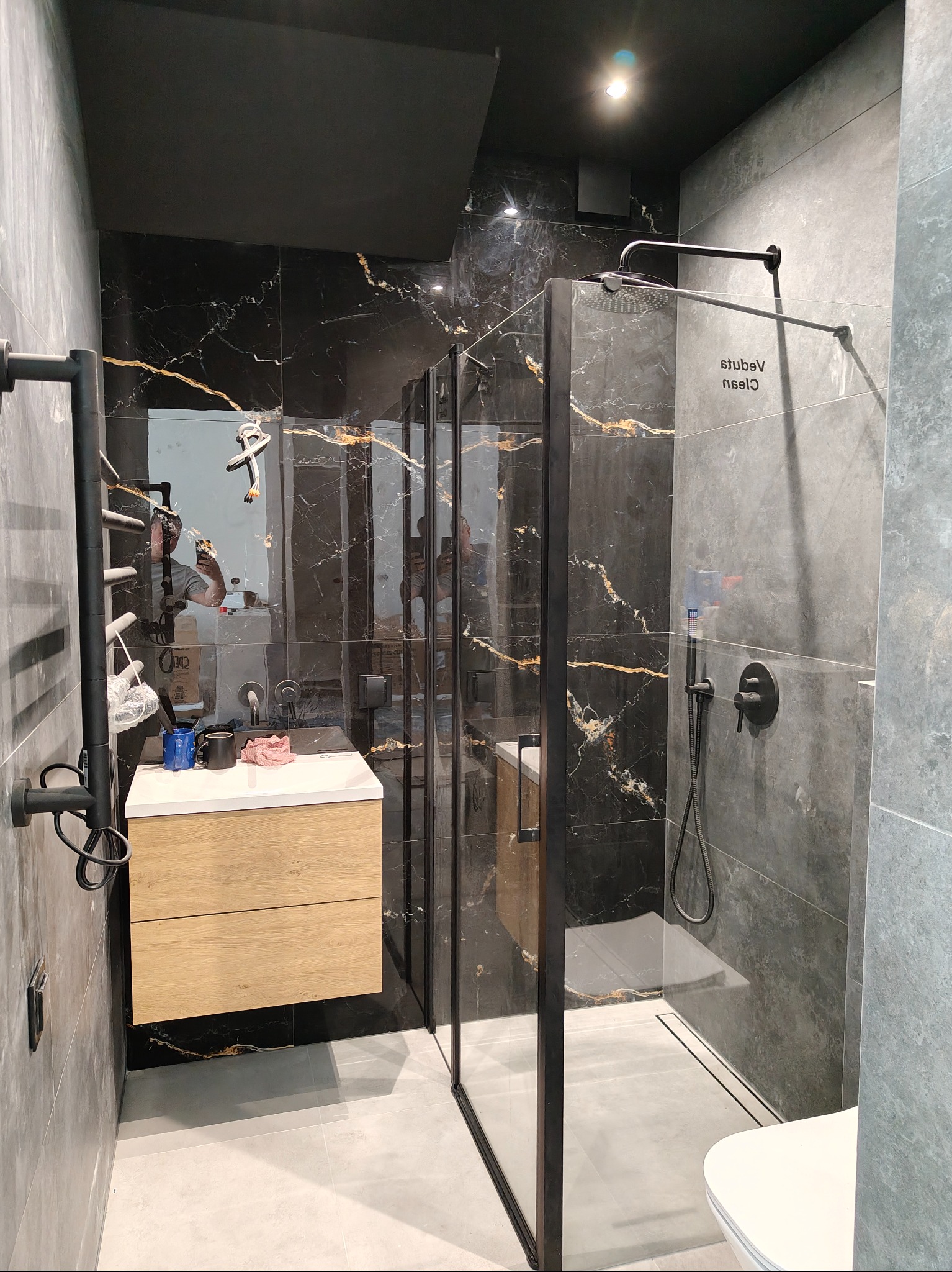 Nowoczesna łazienka z czarnymi płytkami imitującymi marmur, kabiną prysznicową typu walk-in z czarnymi ramami i drewnianą szafką pod umywalką. Widoczny grzejnik drabinkowy.