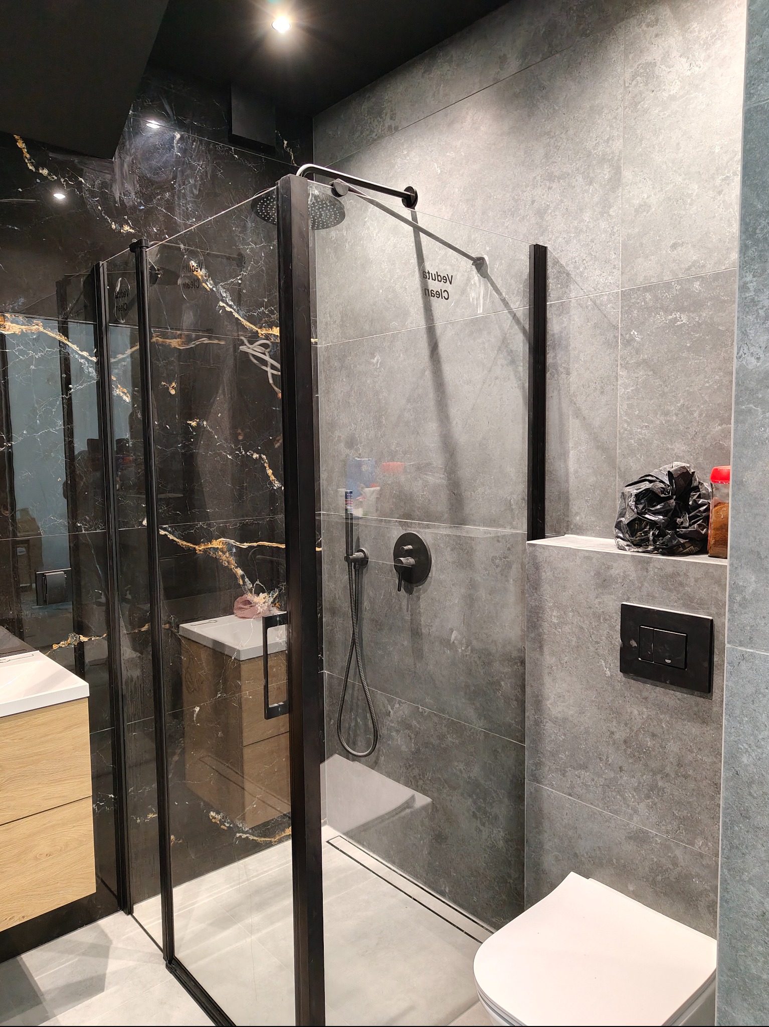 Nowoczesna łazienka z kabiną prysznicową typu walk-in, wykończona płytkami w odcieniach szarości i czerni z elementami złota. Widoczna armatura w kolorze czarnym.