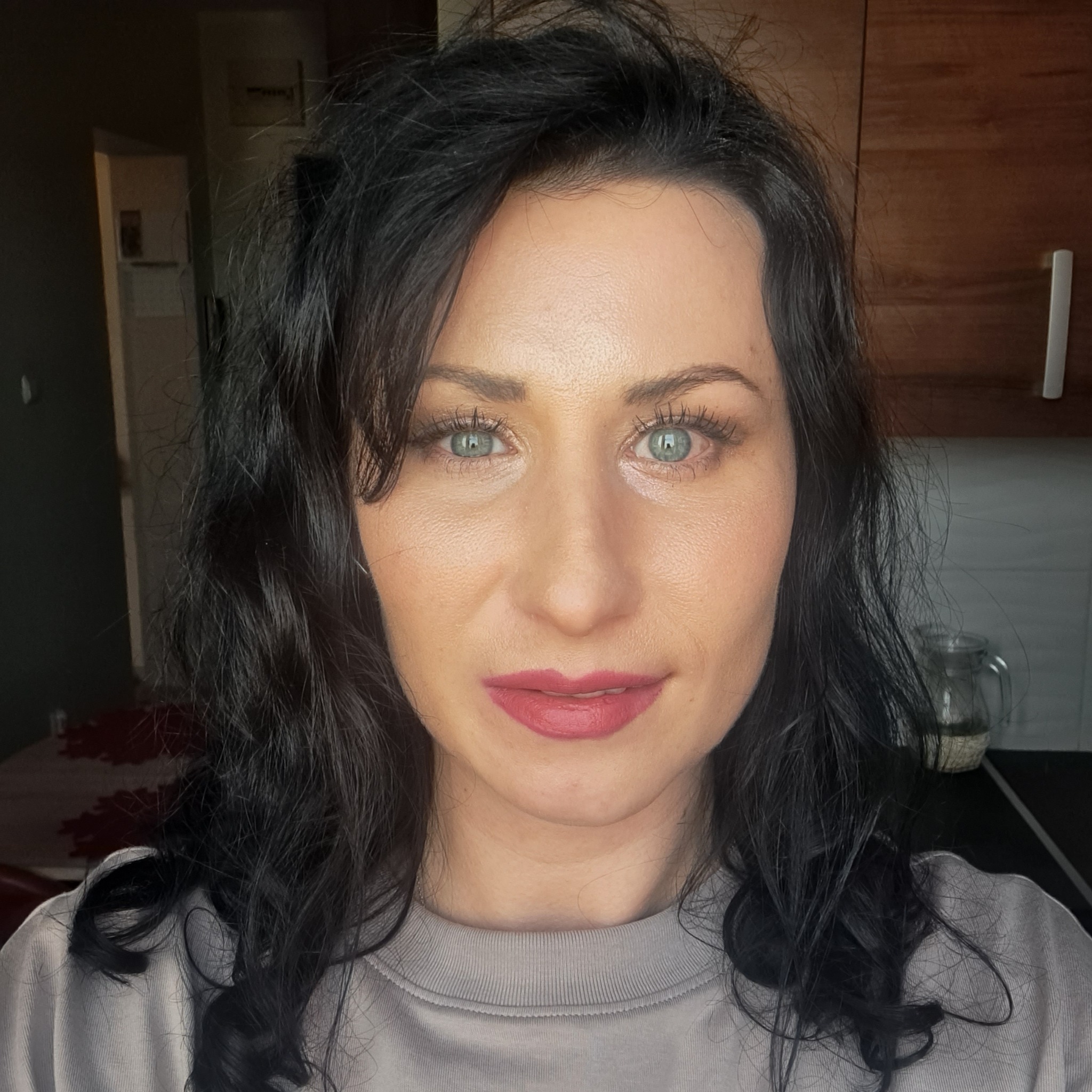 Portret kobiety z ciemnymi, kręconymi włosami i jasnymi oczami, pozującej w pomieszczeniu przypominającym kuchnię, prawdopodobnie prezentującej się jako opiekunka do dziecka.