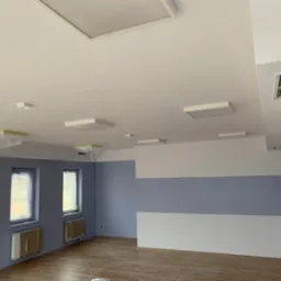 Wnętrze pokoju po malowaniu ścian na szaro i biało, z zabezpieczonymi oknami i grzejnikami folią malarską, widoczne panele podłogowe i nowoczesne oświetlenie sufitowe.