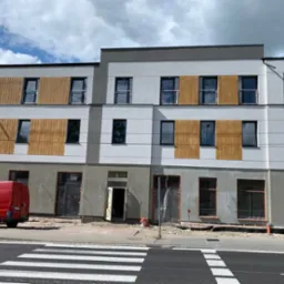 Budynek w trakcie prac elewacyjnych z częściowo zamontowaną drewnianą okładziną, widoczny czerwony samochód dostawczy i fragment przejścia dla pieszych.