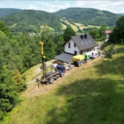 Wiercenie studni głębinowej na zboczu wzgórza obok domu jednorodzinnego, widok z góry na sprzęt wiertniczy i malowniczy krajobraz górski w tle.