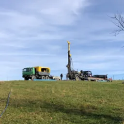 Ciężki sprzęt wiertniczy na zielonym wzgórzu podczas prac wiertniczych, widoczny pracownik w oddali oraz błękitny wąż doprowadzający wodę.