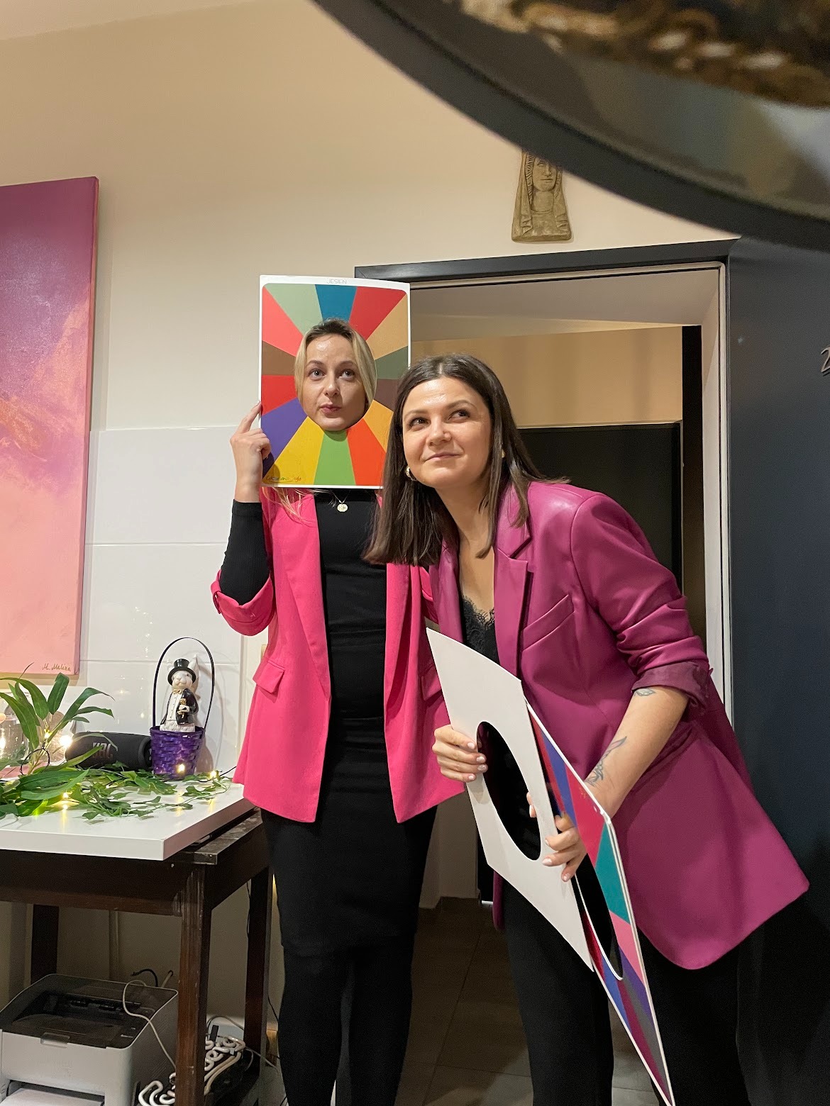Dwie kobiety w różowych marynarkach pozują z rekwizytami do analizy kolorystycznej: jedna trzyma paletę kolorów z wyciętym otworem na twarz, druga trzyma ramkę z próbkami kolorów.