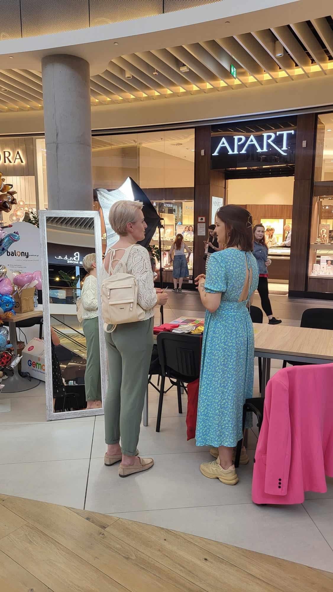 Dwie kobiety rozmawiają w centrum handlowym, jedna w zielonych spodniach i białej bluzce z plecakiem, druga w niebieskiej sukience w kwiaty, w tle sklep Apart.