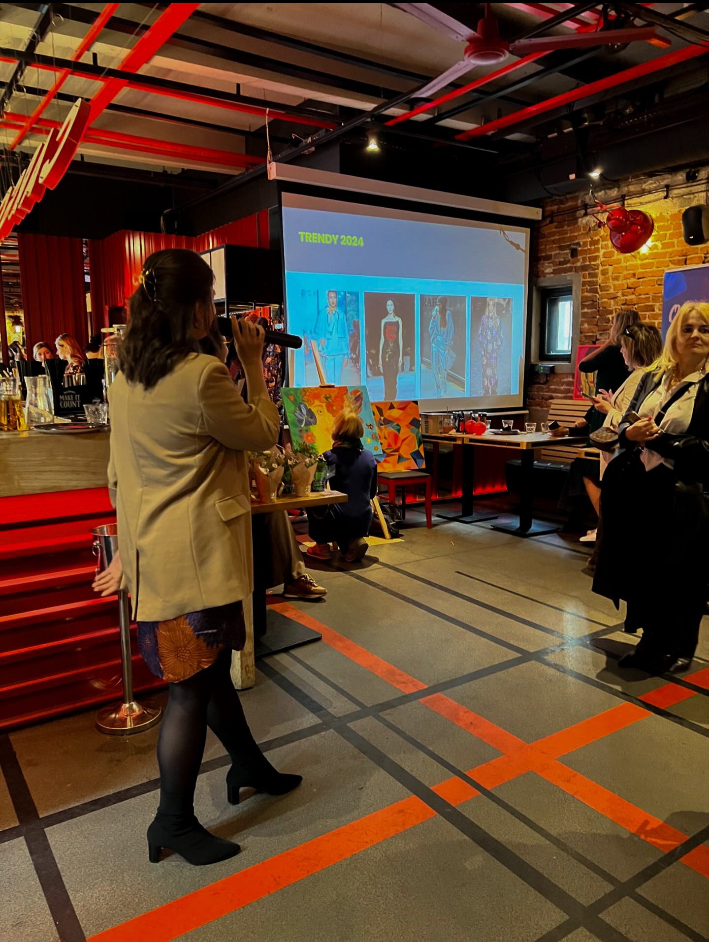 Prezentacja trendów mody 2024: kobieta z mikrofonem na tle ekranu z modelami, publiczność i obrazy na sztalugach.