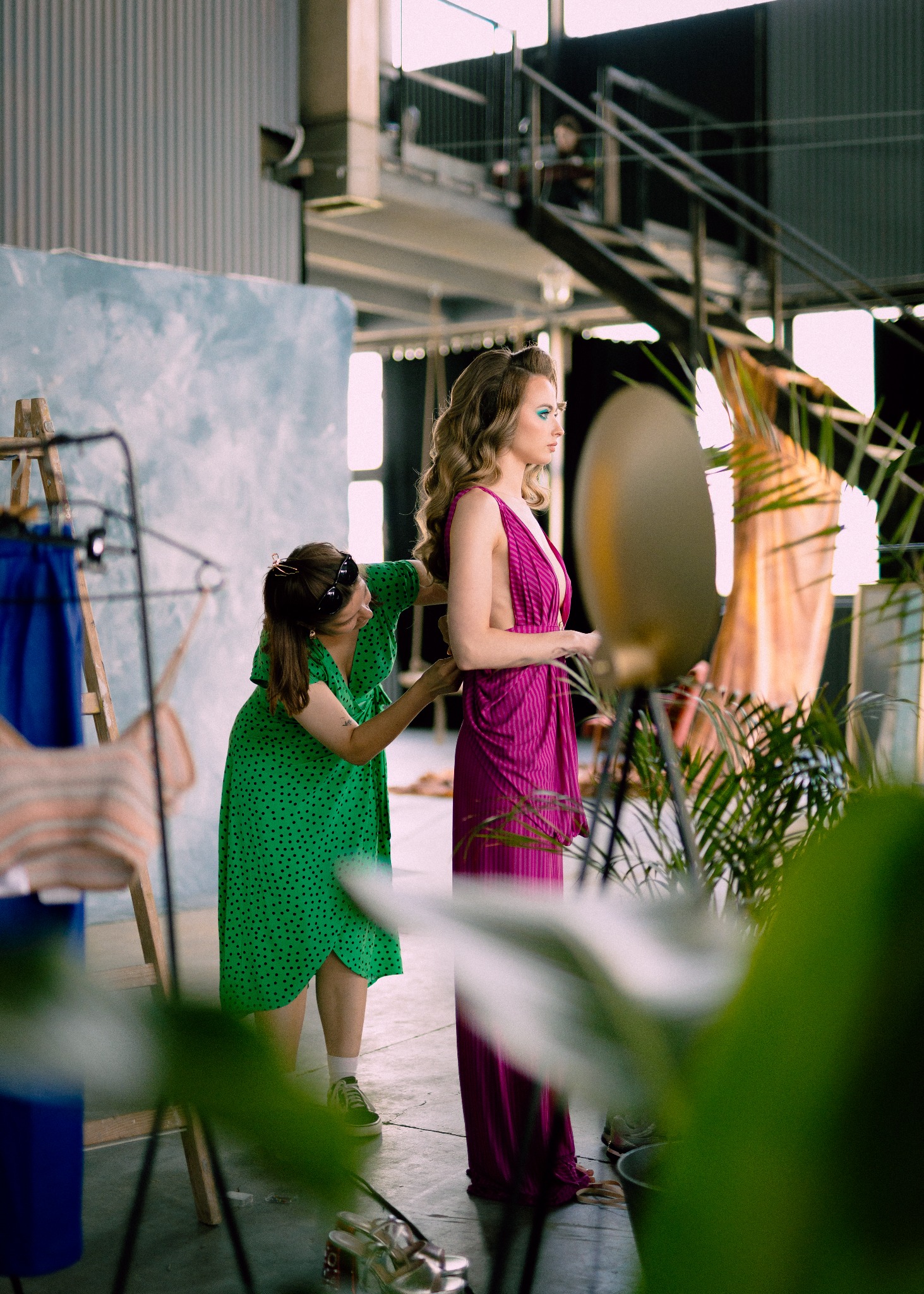 Kobieta w zielonej sukience z czarnymi kropkami dopasowuje fuksjową sukienkę modelce w studio z drabinką, lampą i roślinami w tle.