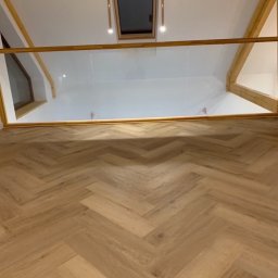 Pikollo - Jasna podłoga ułożona w jodełkę francuską, widok z góry na balustradę ze szkła i drewniane elementy konstrukcyjne poddasza z oknem dachowym i wiszącymi lampami.