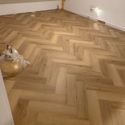 Pikollo - Ukończone podłogi z paneli ułożonych w jodełkę w pokoju na poddaszu, widoczne listwy przypodłogowe i wystający kabel elektryczny.