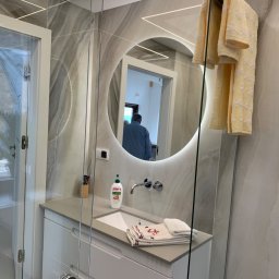 Pikollo - Nowoczesna łazienka z okrągłym lustrem podświetlanym LED, szklaną kabiną prysznicową i umywalką z szafką w kolorze białym, widoczne ręczniki z motywem marynistycznym.