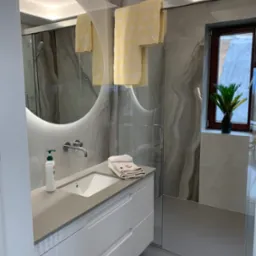 Nowoczesna łazienka z podświetlanym okrągłym lustrem, umywalką nablatową, białymi szafkami pod umywalką, ręcznikami w kolorze żółtym i szarymi płytkami imitującymi marmur na ścianach.