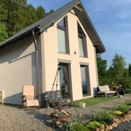 Nowoczesny dom z jasną elewacją, szarym dachem i ramami okiennymi, otoczony ogrodem z paleniskiem tr&oacute;jnożnym, drewnianą ławką z palet i roślinami, w tle las i quad.