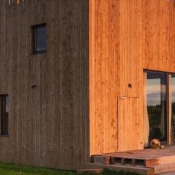Pikollo - Nowoczesna elewacja budynku z drewnianymi elementami w Jaszkówce, widoczna z zewnątrz, z unikalnym pionowym układem desek na szczycie i ciepłym oświetleniem słonecznym.