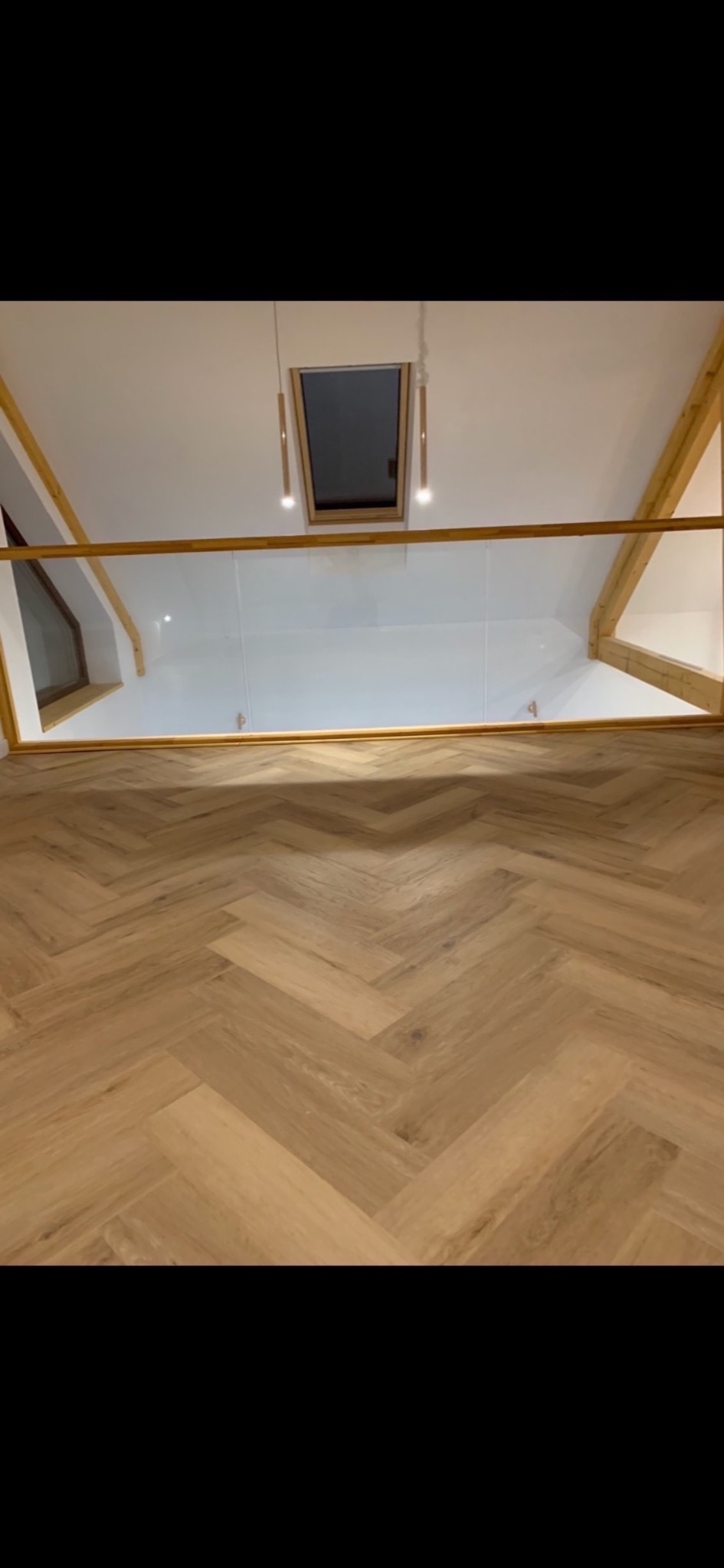 Jasna podłoga ułożona w jodełkę francuską, widok z góry na balustradę ze szkła i drewniane elementy konstrukcyjne poddasza z oknem dachowym i wiszącymi lampami.