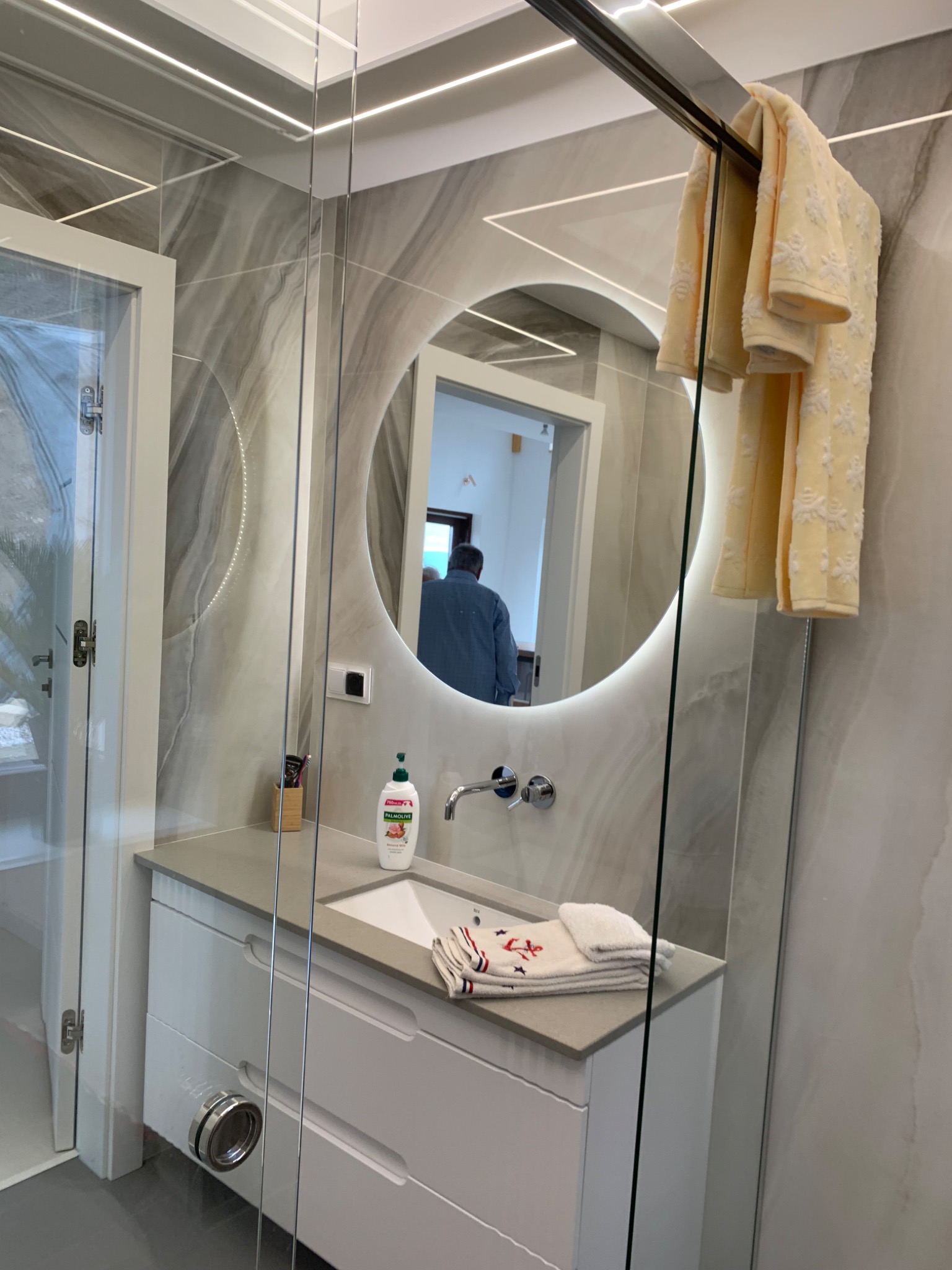 Nowoczesna łazienka z okrągłym lustrem podświetlanym LED, szklaną kabiną prysznicową i umywalką z szafką w kolorze białym, widoczne ręczniki z motywem marynistycznym.