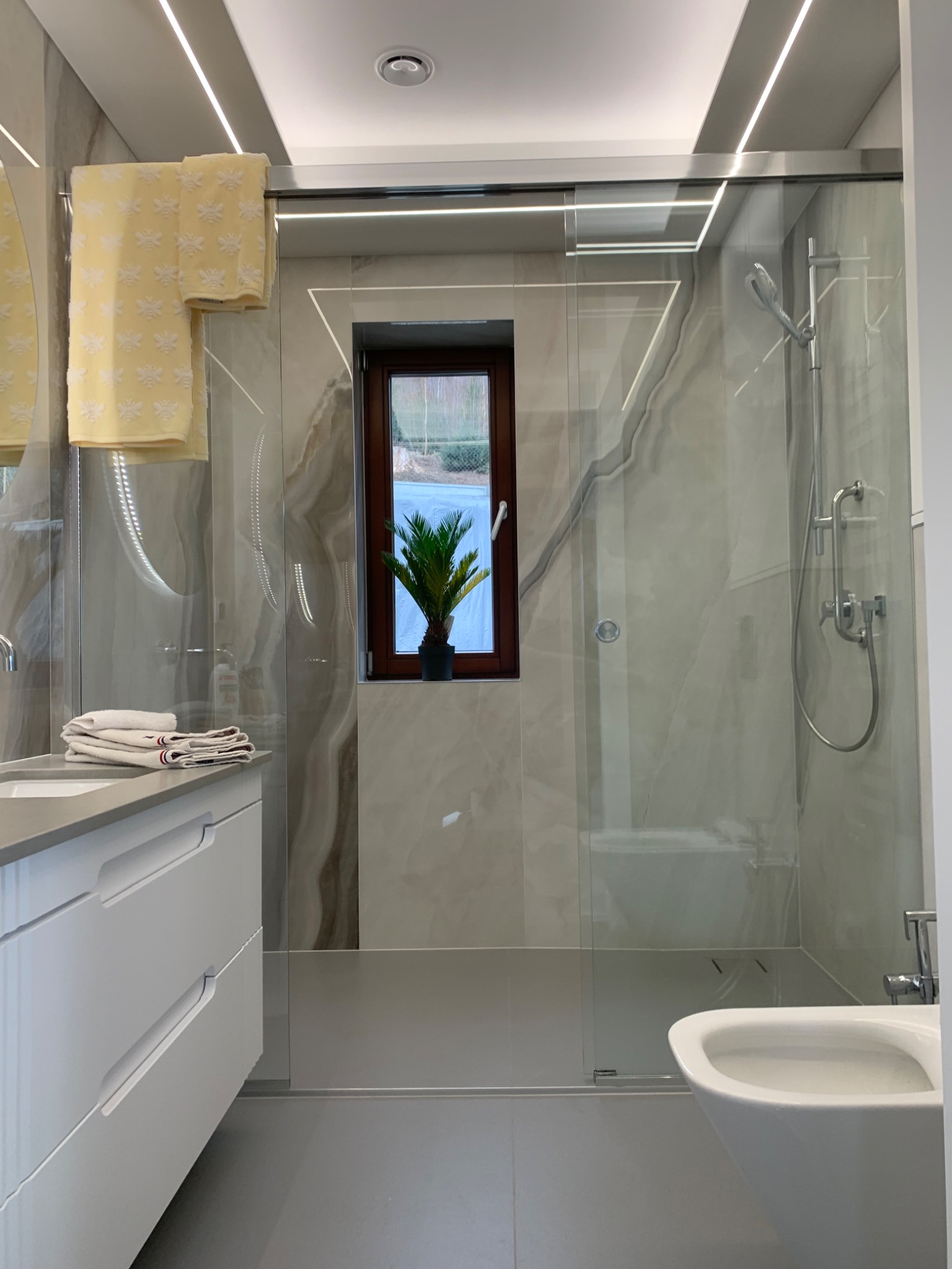 Nowoczesna łazienka z kabiną prysznicową typu walk-in, marmurowymi płytkami, białymi meblami łazienkowymi i oknem z rośliną doniczkową.