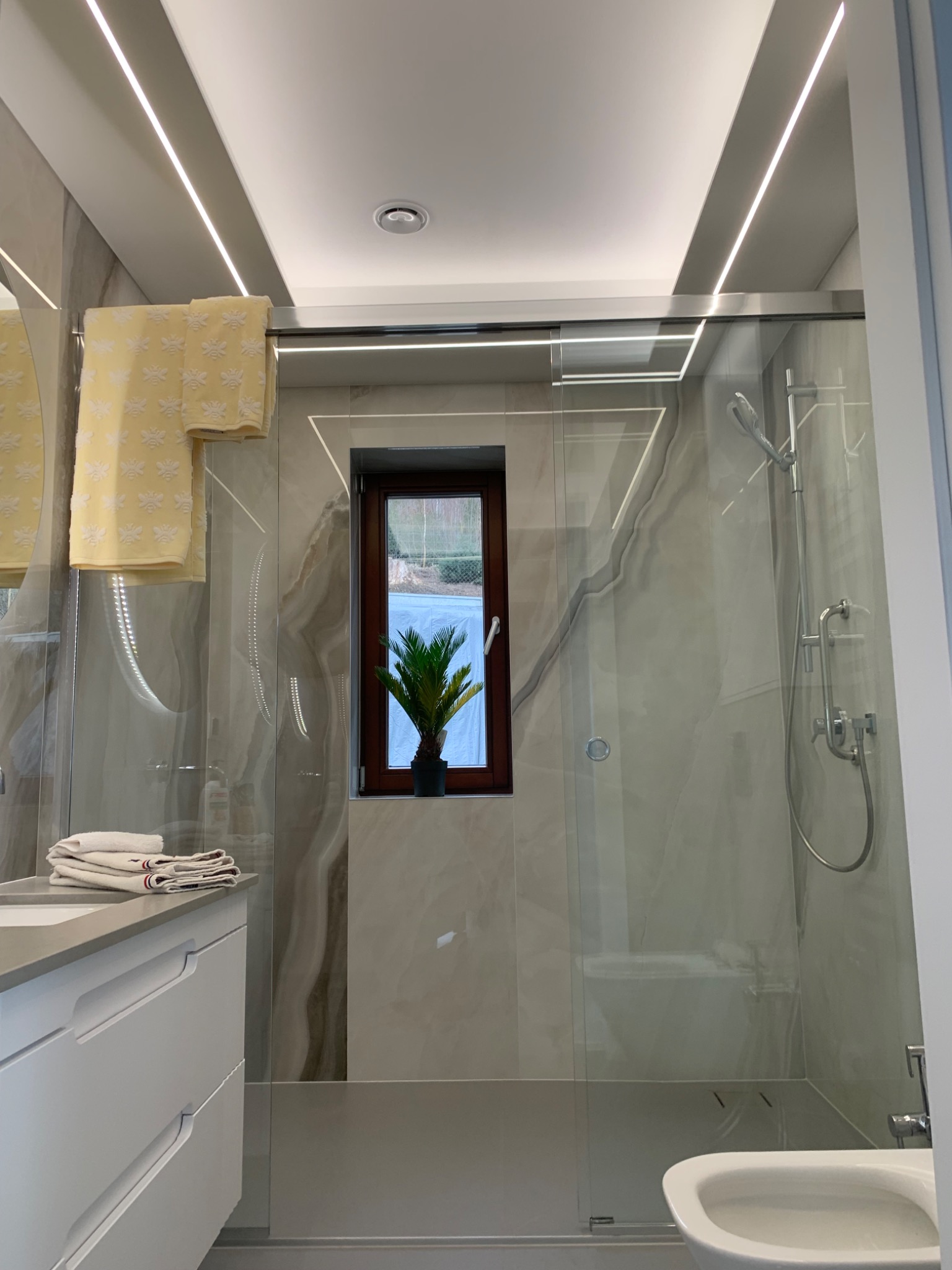 Nowoczesna łazienka z marmurowymi płytkami, szklaną kabiną prysznicową, podświetleniem LED, oknem z widokiem na zieleń i rośliną doniczkową.