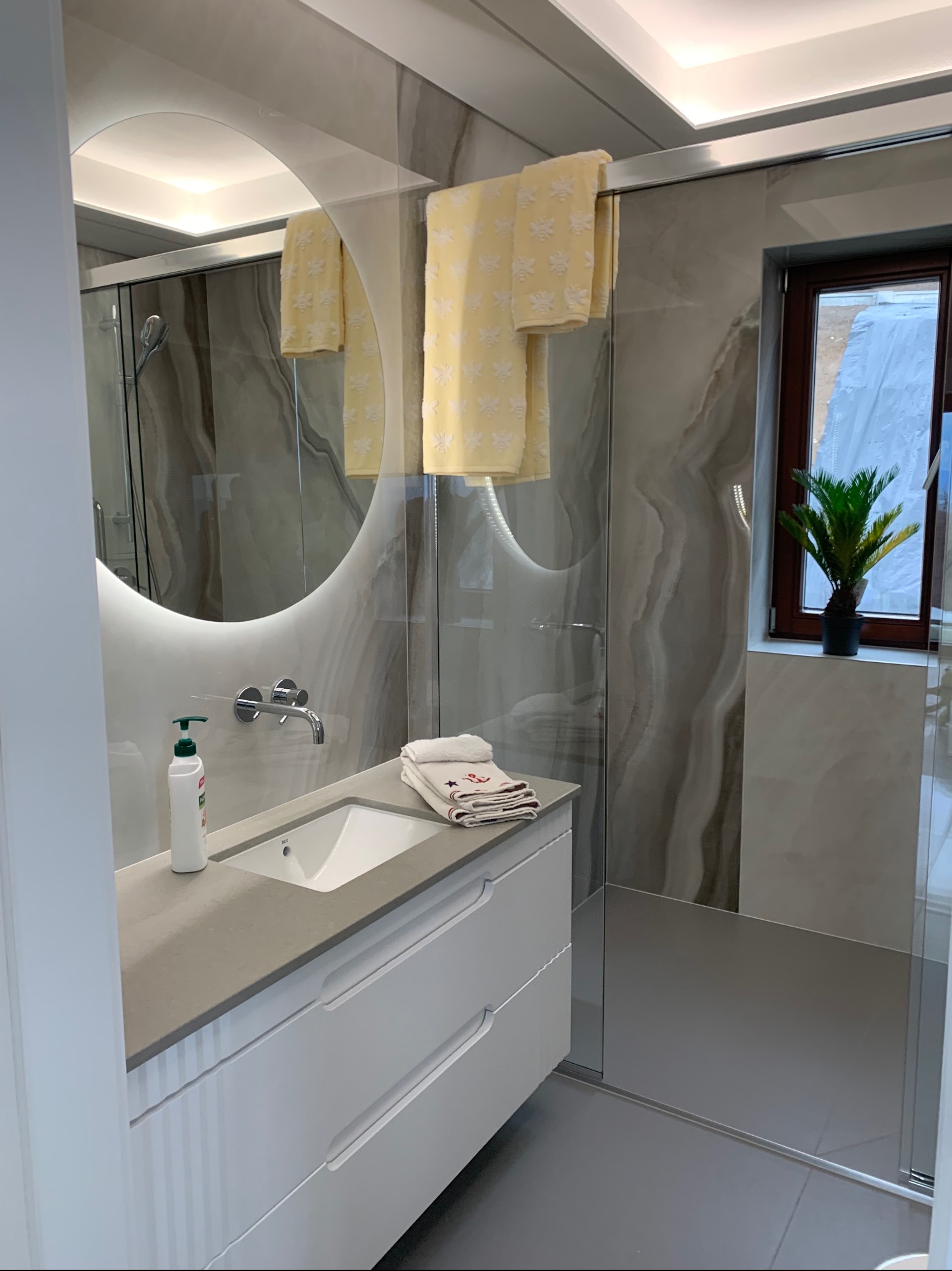 Nowoczesna łazienka z podświetlanym okrągłym lustrem, umywalką nablatową, białymi szafkami pod umywalką, ręcznikami w kolorze żółtym i szarymi płytkami imitującymi marmur na ścianach.