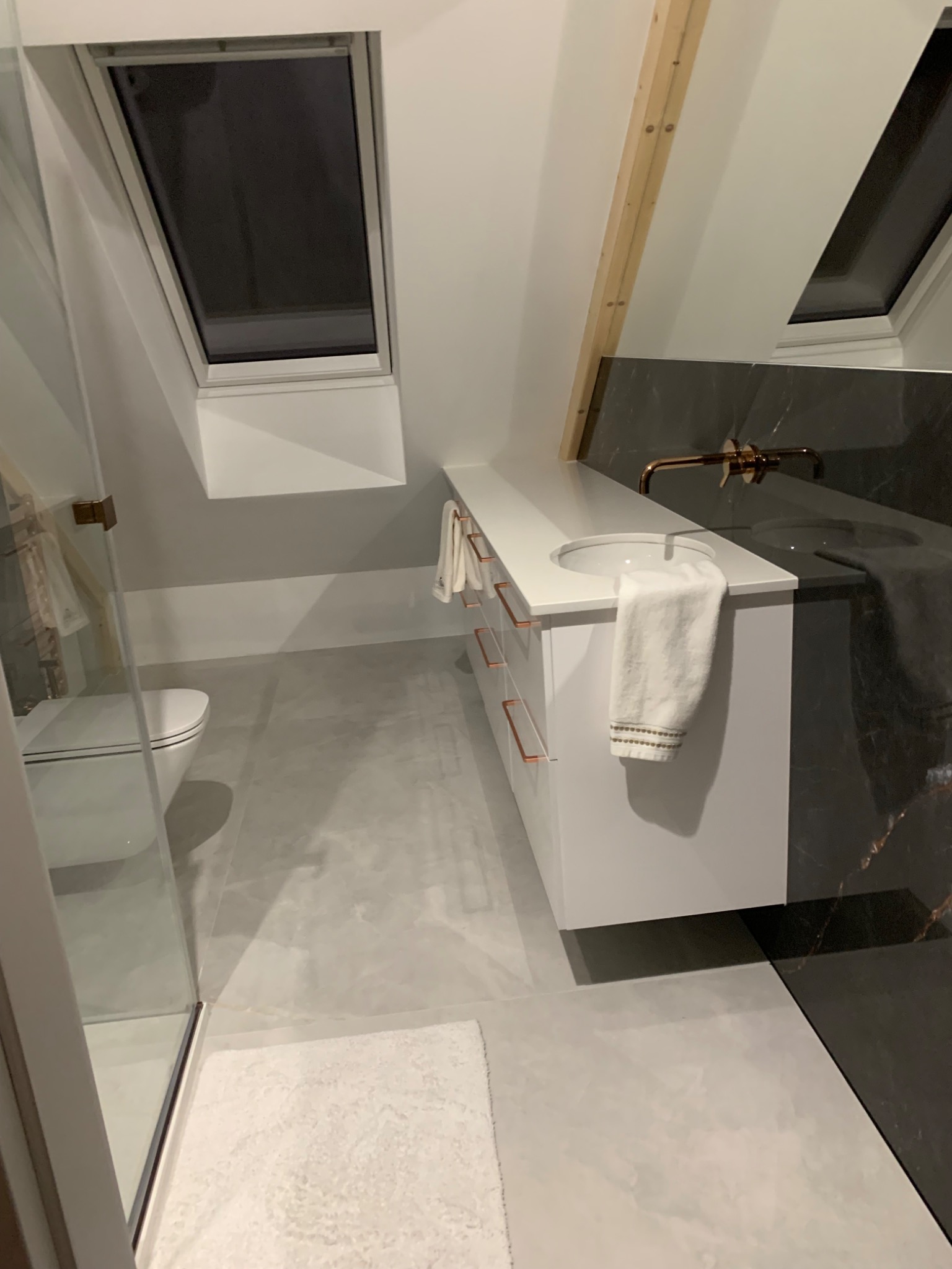 Nowoczesna łazienka na poddaszu z oknami dachowymi, białą szafką z umywalką i złotą armaturą na tle czarnego marmuru, widoczna kabina prysznicowa i toaleta.