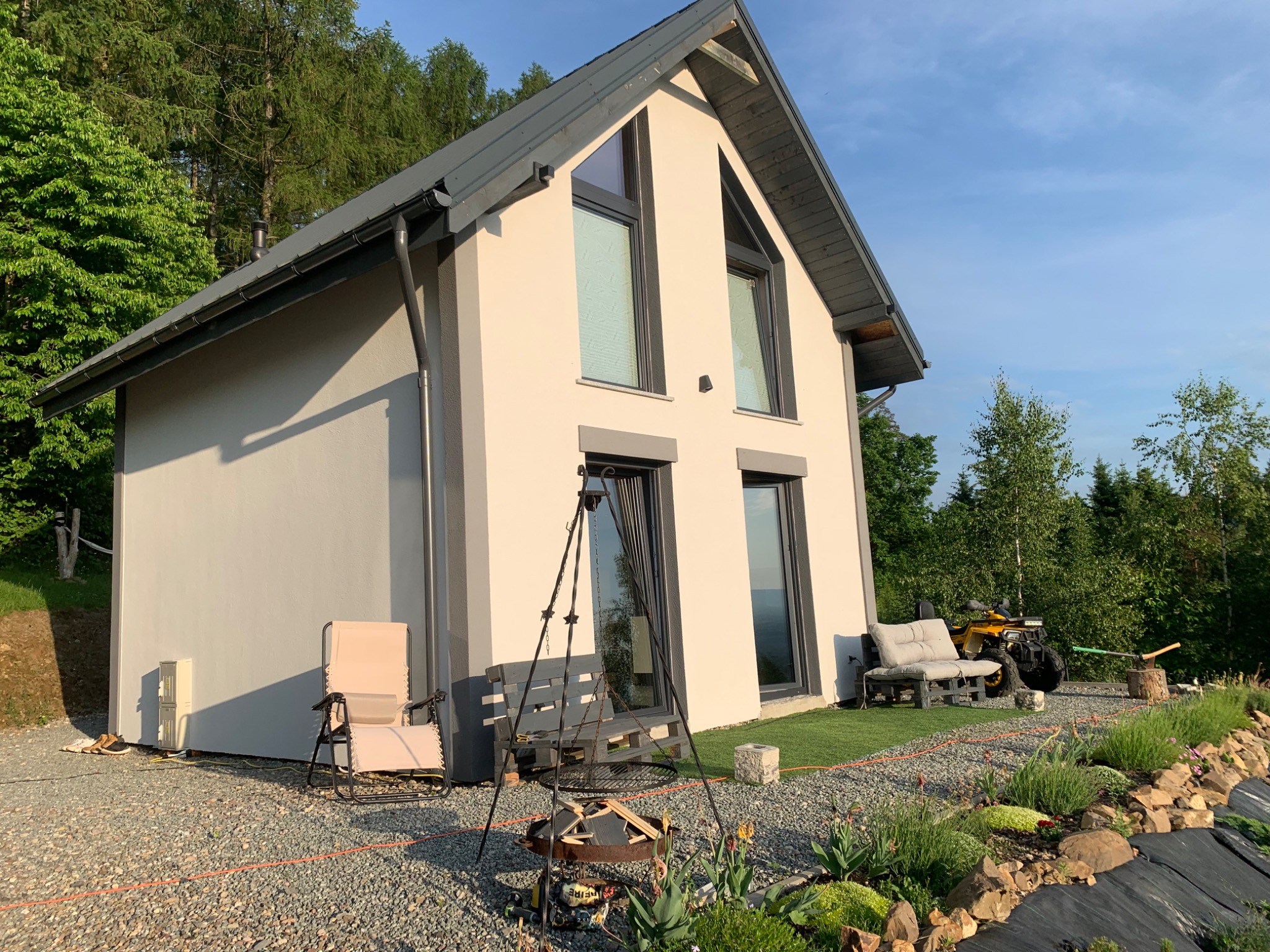 Nowoczesny dom z jasną elewacją, szarym dachem i ramami okiennymi, otoczony ogrodem z paleniskiem trójnożnym, drewnianą ławką z palet i roślinami, w tle las i quad.