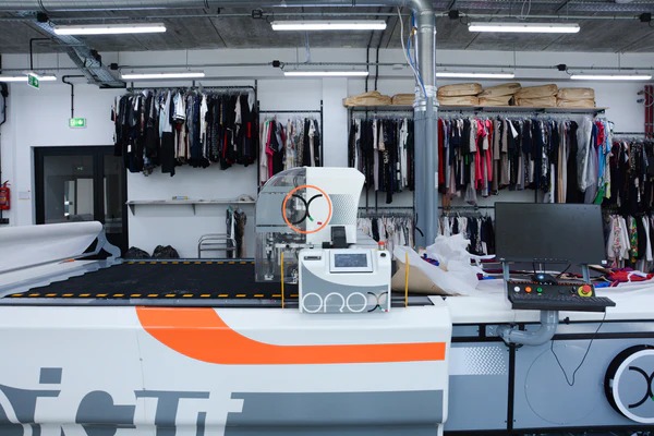 Automatyczna krajalnica tkanin z pomarańczowym akcentem w nowoczesnej, dobrze oświetlonej pracowni odzieżowej. W tle wieszaki z ubraniami.