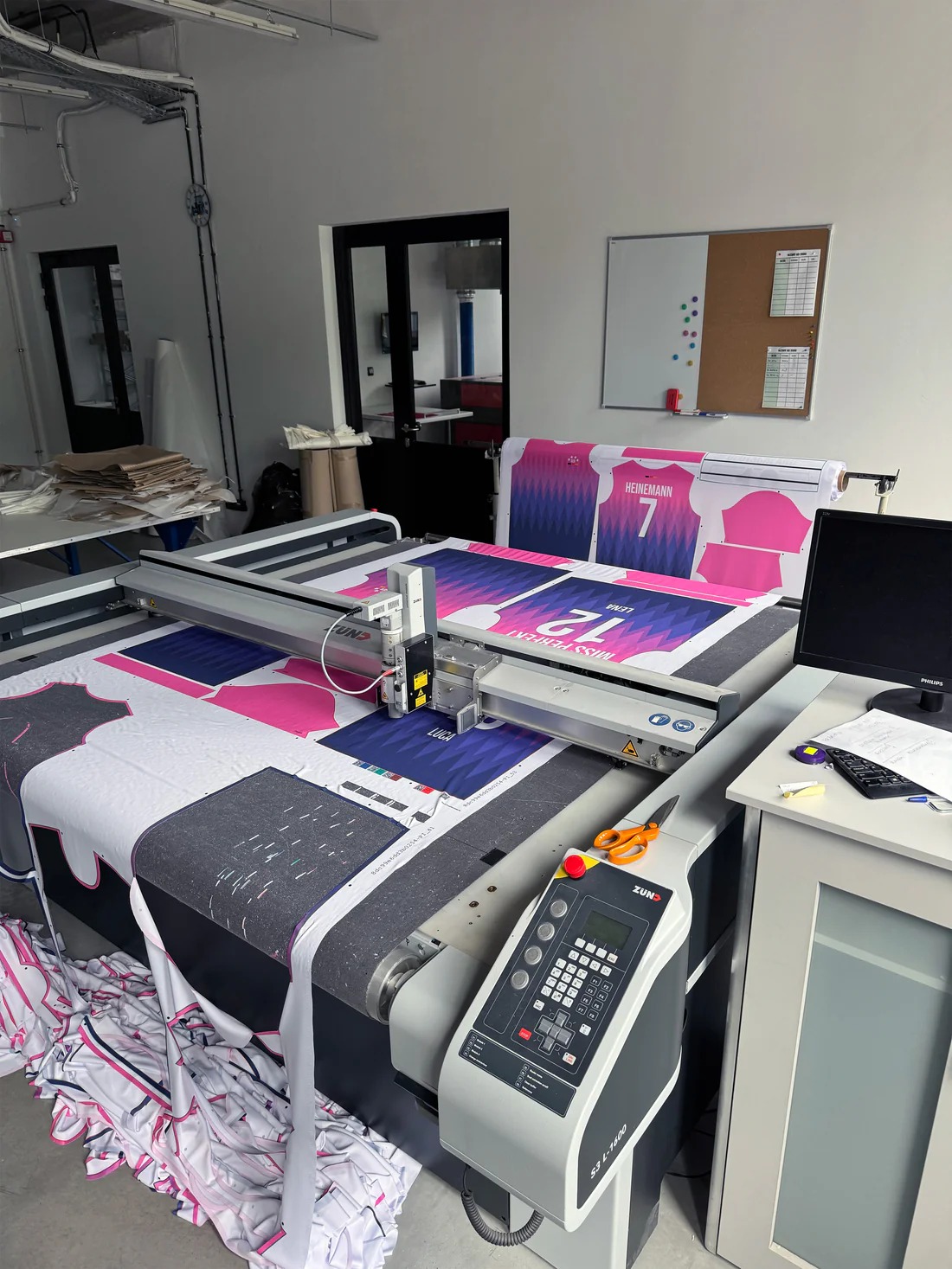 Automatyczna krojownia Zund S3 L-1600 tnąca materiał na koszulki sportowe z nadrukiem, widoczne nożyczki i monitor obok maszyny.