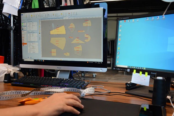Projektowanie wykrojów odzieży na komputerze: widok ekranu z programem CAD/CAM, obok miarka krawiecka, nożyczki i dłoń obsługująca mysz, drugi monitor w tle.