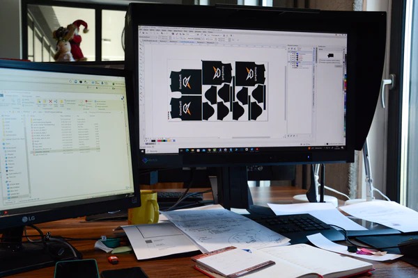 Projekt wykroju odzieży z logo na ekranie komputera, widok z biurka z notatkami i kubkiem.