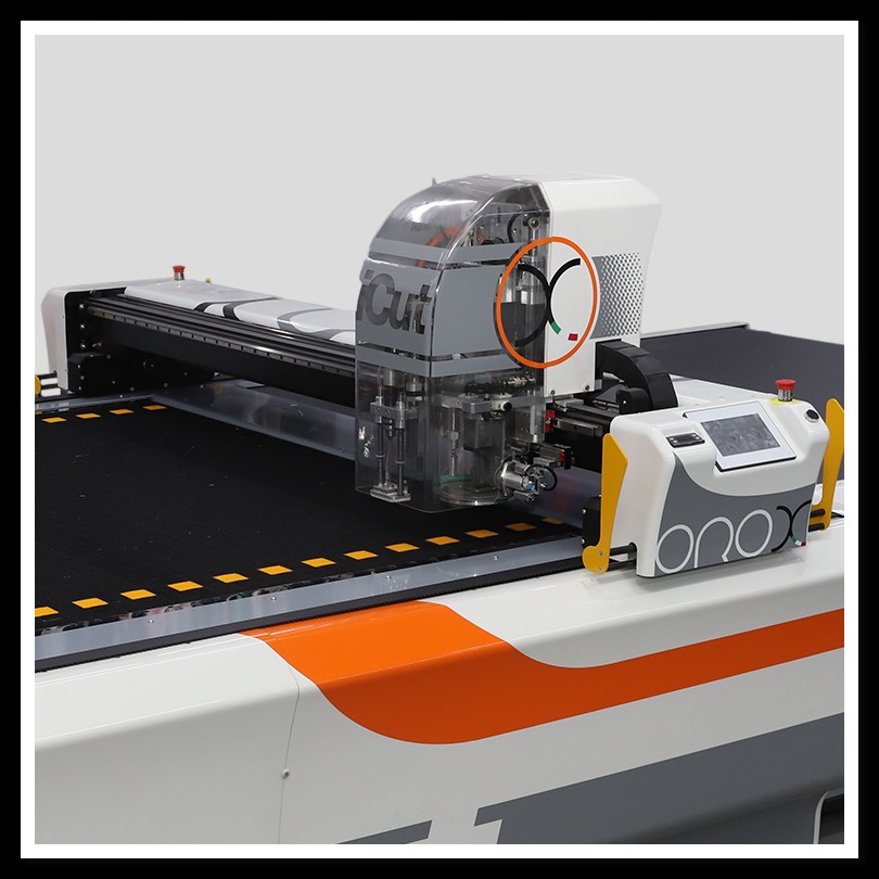 Automatyczny ploter tnący z przezroczystą osłoną, widoczne ostrze tnące, panel sterowania z ekranem dotykowym i pomarańczowe akcenty na białej obudowie.