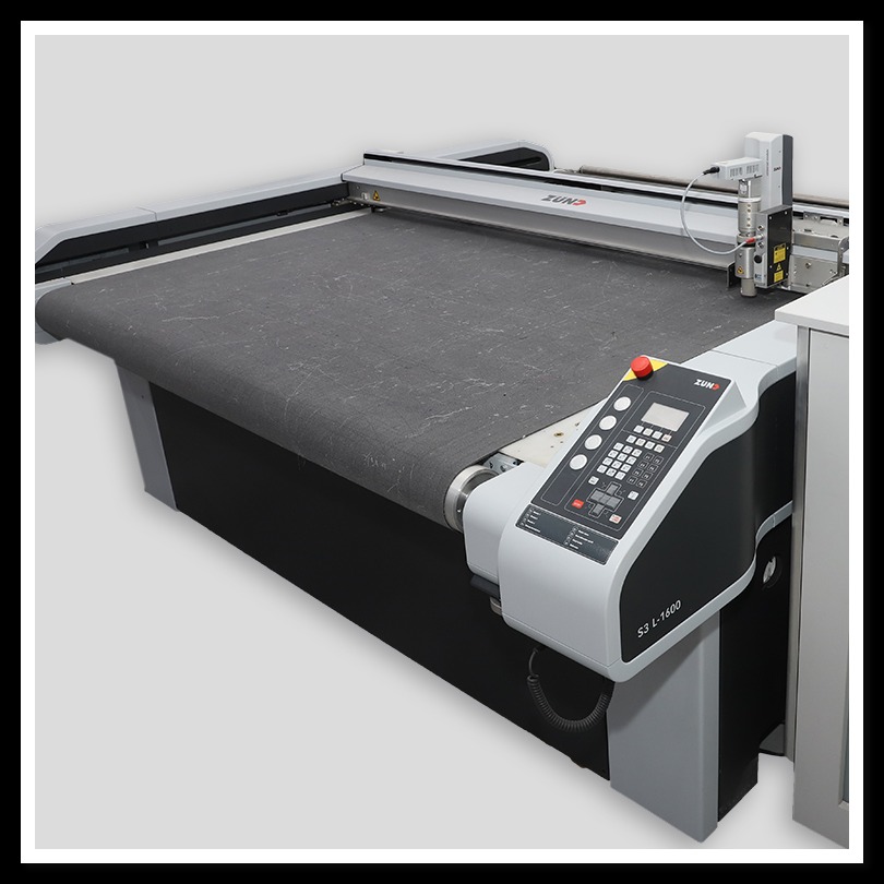 Profesjonalna maszyna tnąca Zund S3 L-1600 z szarym materiałem na stole roboczym, widoczny panel sterowania z przyciskiem awaryjnym.