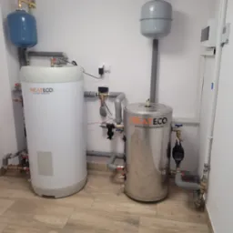 Dwa zbiorniki systemów grzewczych marki HEATECO, jeden biały, drugi ze stali nierdzewnej, z podłączonymi rurami i osprzętem, zainstalowane w pomieszczeniu z płytkami imitującymi drewno.