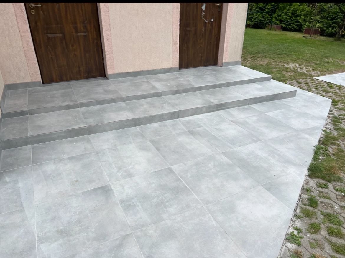 Wykończone płytkami schody wejściowe do budynku w Wólce Kosowskiej. Szare płytki ułożone na stopniach i przed wejściem, widoczne drzwi wejściowe.