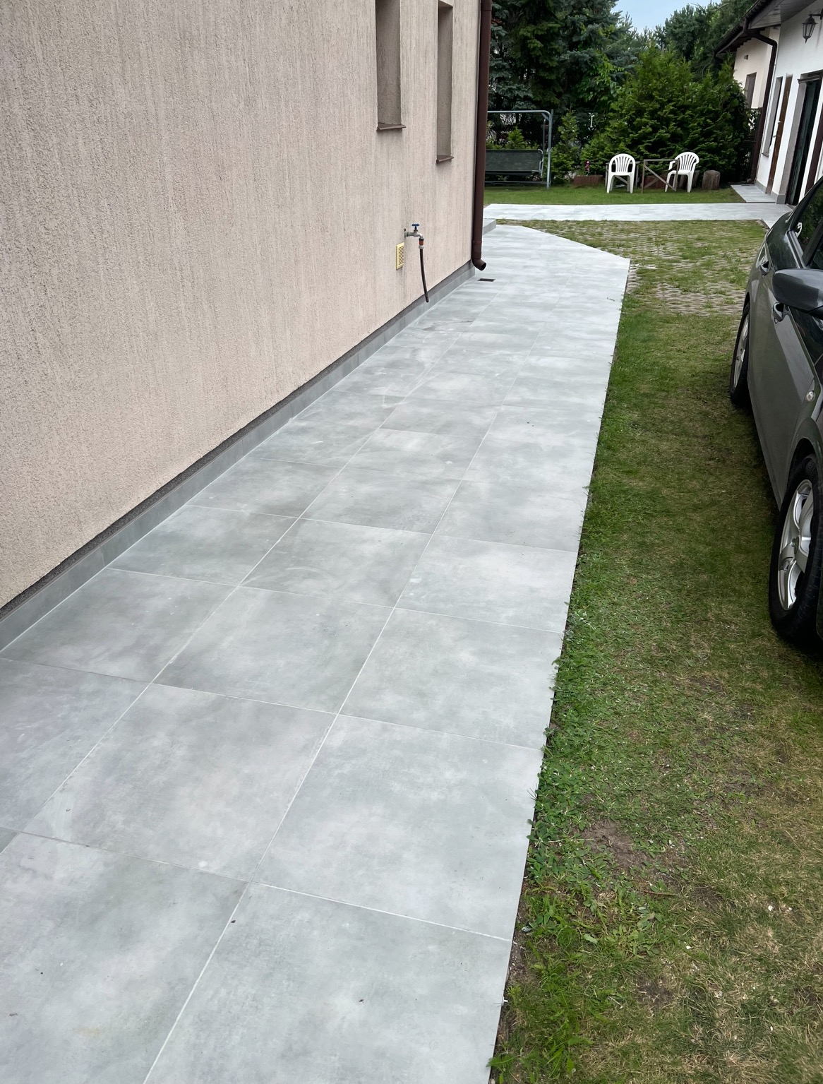 Nowoczesna, szara ścieżka z płytek ułożona wzdłuż elewacji domu, z widocznym fragmentem trawnika i samochodu. Minimalistyczny design.