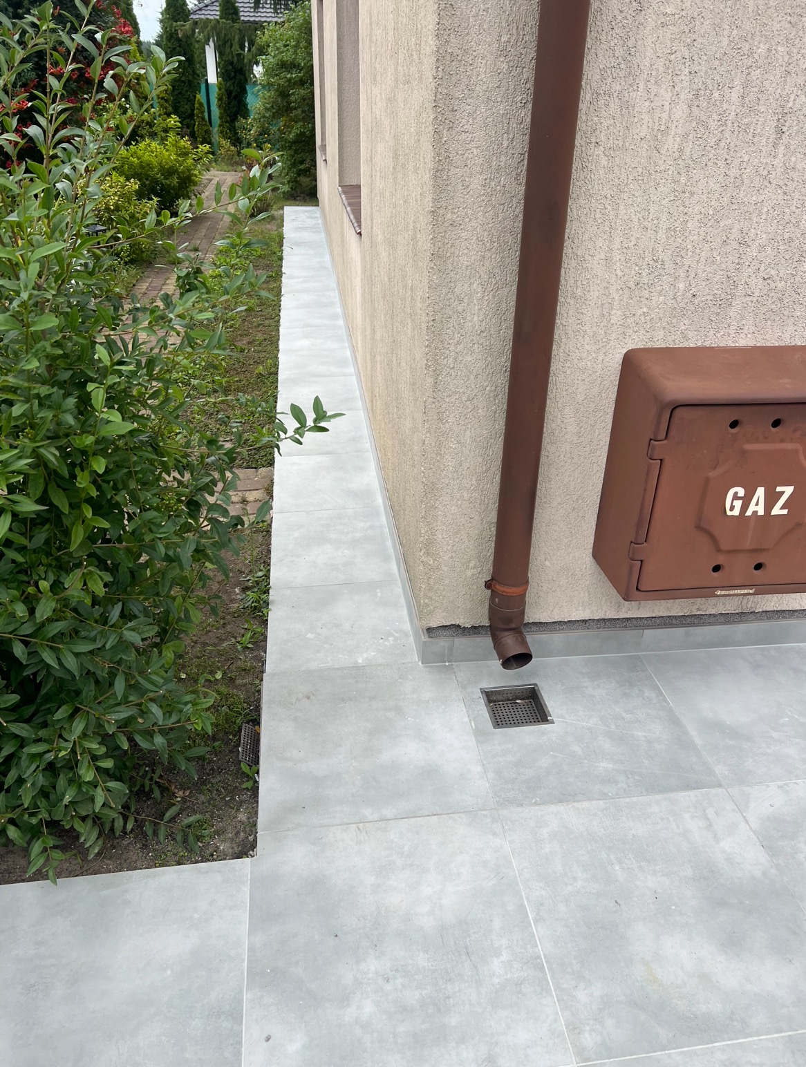 Szare płytki tarasowe ułożone wzdłuż ściany budynku, z brązową rynną i skrzynką gazową. Widoczny fragment zieleni. Eleganckie wykończenie przestrzeni.