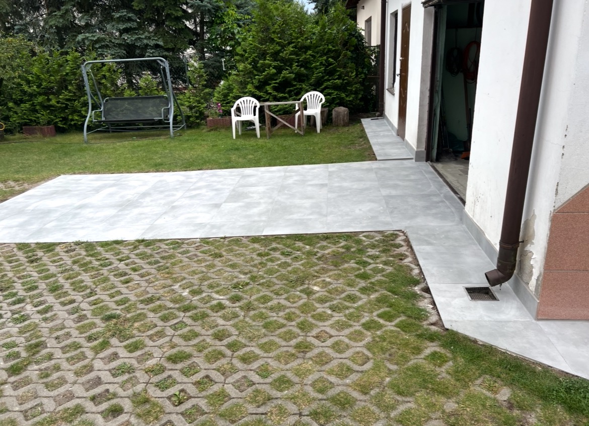 Ułożona, szara, kwadratowa terakota na zewnątrz budynku, częściowo na trawie i kostce ażurowej, prowadząca do drzwi. W tle huśtawka i krzesła.